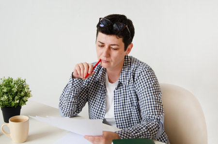 A middle-aged woman sitting at a table in the office holds documents in her hand. woman makes notes, studying papers. concept of home office, learning, planningの写真素材