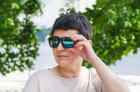 Portrait of a middle-aged woman with black short hair and sunglasses. A woman looks into the distance, standing against the backdrop of trees on a sunny summer dayの写真素材