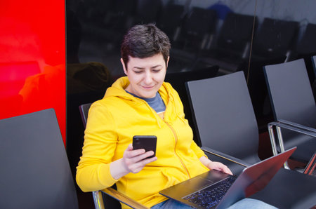 female traveler working on a laptop and using a smartphone while sitting in the airport terminal...