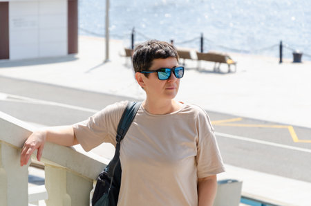 Portrait of middle-aged woman with black short hair and sunglasses. A woman looks into the distance, standing against the backdrop of the city embankment on a sunny summer dayの写真素材
