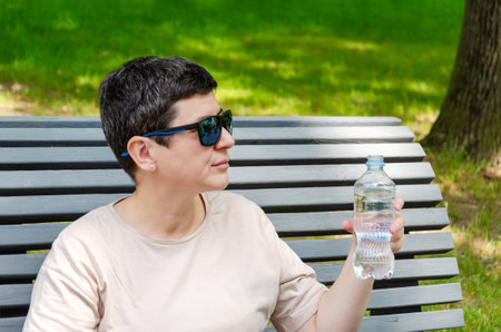 middle-aged woman with black short hair and sunglasses drinks water from a bottle on a hot summer day, sitting on a bench in the parkの写真素材