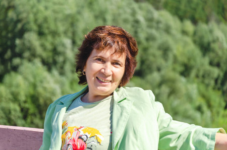 Attractive smiling elderly woman stands on the bridge on a summer sunny day against the backdrop of...