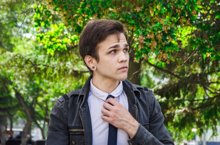 Portrait of a stylish young man sitting on a park bench. An attractive guy in a shirt and jacket straightens his necktieの写真素材