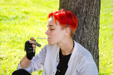 Portrait of a red-haired young woman smoking a cigarette. a girl with a short haircut with a cigarette in her hands sits on the lawn in the parkの写真素材