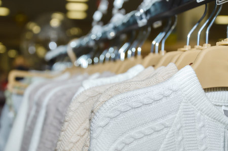 Rack in a modern children's clothing store with white and beige knitted cotton sweaters on hangers,...