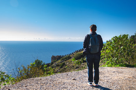 human stands on a cliff with a backpack, gazing out over a vast ocean and picturesque coastline. The...