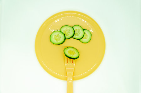 Fresh cucumber slices neatly arranged on a vibrant yellow plate, with a single slice resting on a matching fork, showcasing a minimalist and healthy food presentation.の写真素材