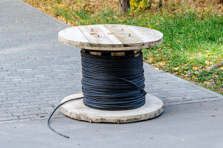 Large wooden spool wound with black cable sits on a sidewalk, surrounded by grass and autumn leaves. Industrial and construction materials are showcased in an outdoor setting.の写真素材