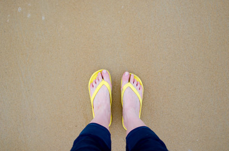 Feet wearing yellow flip flops stand firmly on soft sandy beach. The image captures a moment of...