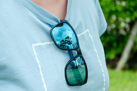 Close-up of sunglasses hanging on collar a shirt, reflecting palm trees and a building. The...