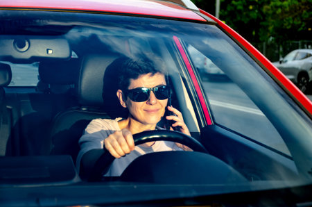 Woman wearing sunglasses drives a red car while talking on a phone. Captured through the windshield,...