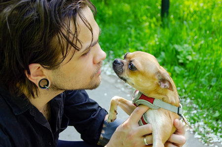 man kneels on a path holding his small dog, hugging and kissing it. The dog is in a harness, and they engage in a close, affectionate interaction amidst green surroundings.の写真素材