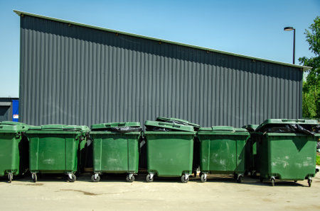 Industrial setting with several green trash bins lined up against a large corrugated metal wall. The bins are intended for waste disposal, emphasizing cleanliness and organization.の写真素材