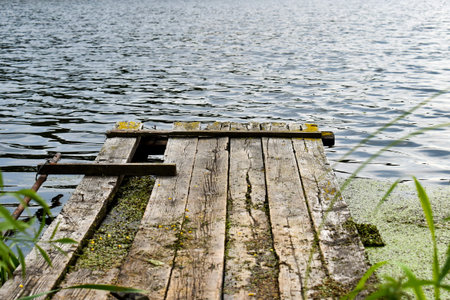 Weathered wooden pier extending into calm lake water with gentle ripples. The old dock shows moss,...