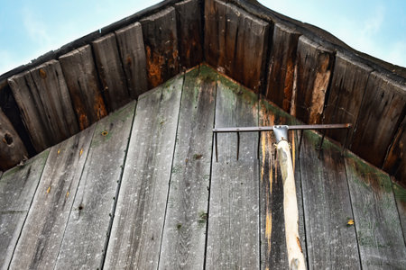 A vintage wooden rake rests against the gray weathered wall of an old wooden barn. The rustic wood texture and aged tool evoke a countryside farming atmosphere under a blue sky.の写真素材