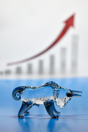 A transparent glass bull figurine standing on a blue surface with a blurred background of a bar chart and red upward arrow, symbolizing a bullish stock market trend and economic growth.の写真素材