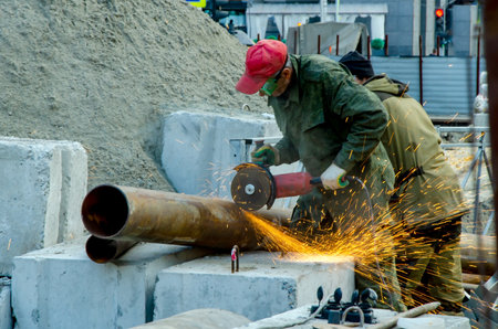 Man in protective clothing large metal pipe with angle grinder outdoors, bright sparks flying during industrial construction work.の写真素材