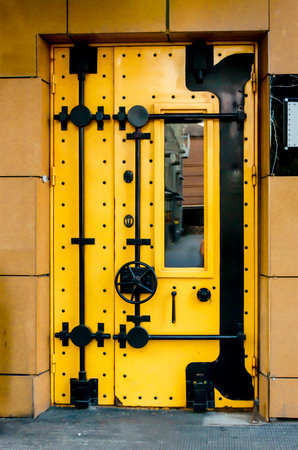 Bright yellow metal door with black bolts and lock hardware, designed to resemble a bank vault. Installed as entrance in building facadeの写真素材