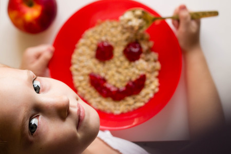 little girl eating appetizingly oatmeal from a red plate with a smiley of jam, healthy food, breakfast, childrens menuの写真素材