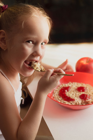 little girl eating appetizingly oatmeal from a red plate with a smiley of jam, healthy food, breakfast, childrens menuの写真素材