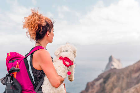 Curly hair woman is holding her maltese dog in the arms and happily smiling on the mountain topの写真素材