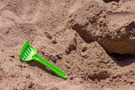 Kids toy rake lost among yellow sand of beach. Green plastic rake closeup on sea sand textured background. Summer beach vacation backdropの写真素材