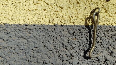 Rusty metal hook of obsolete latch hanging on rough stone wall background of yellow and grey stripes. Texture of uneven plaster wall and old haspの写真素材