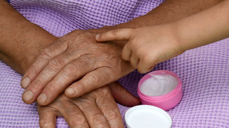 Small girl take cosmetic cream on her finger and apply to granny wrinkled skin hand. Diverse hands...