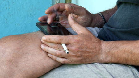 Asian male hands hold cigarette and tap smartphone screen on laps. Man smokes and touches mobile device while smokingの写真素材