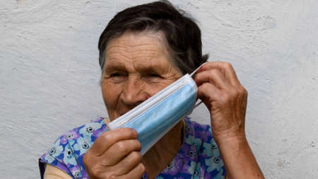 Elderly wrinkled woman putting on protective medical face mask. Old lady shows how to wear mask in appropriate way. Senior people protection while lockdownの写真素材
