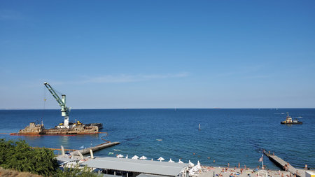 Wrecked cargo ship lies on side near coastline with sandy beach. Floating crane near barrier of...