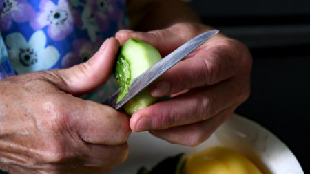 Elder person hands peel of raw green cucumber with kitchen knife. Old wrinkled hands prepare fresh vegetable for veggie salad. Authentic rural real life sceneの写真素材