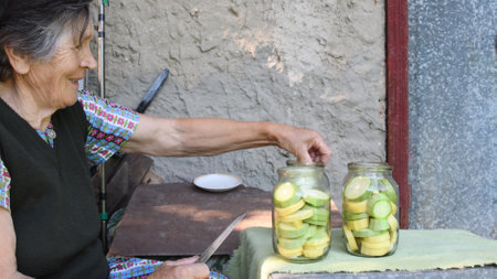 Wrinkled senior Asian woman smile and put sliced raw squash into jar for homemade canning. Rural local cuisine with organic farming vegetables. Zucchini preparation for preservationの写真素材