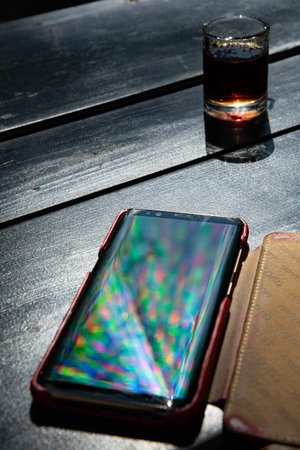 Vibrant rainbow rays reflect in mobile phone screen on black table with alcohol drink shot glass....