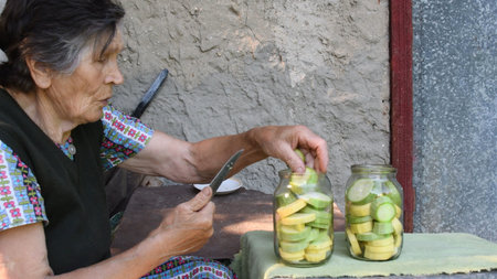 80s mixed race senior woman place zucchini slices into jars as preservation. Granny make homemade...