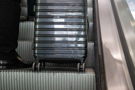 Stylish grey suitcase with wheels on escalator moving stairs near businessman leg in expensive suit and shoes. Business trip concept. Closeup reflective surface of modern hand luggage in airportの写真素材