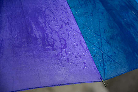 Edge of wet waterproof textile surface of blue and purple umbrella under drizzle rain. Water drops dripping at blue umbrella fabric texture with metal spoke. Rainy weather abstract backgroundの写真素材