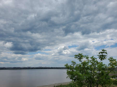 High overcast sky with white fluffy cumulus clouds floating in wind. Tranquil cloudscape over calm water. Altocumulus and cirrocumulus types of clouds and lake shore with flowering elderberry bushの写真素材