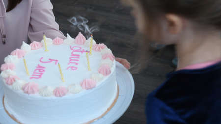 Toddler girl blows out candles at cake on her birthday. Girly white cake with candles and pink cream text - Alice 5 years old. Child looks at cake and makes birthday wish. Authentic happiness conceptの写真素材