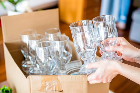 Female hands packing fragile glassware with care using bubble wrap. Put fragile glasses into cardboard box for movingの素材