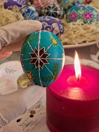 Expert artisans carefully create turquoise Easter egg featuring traditional Ukrainian pysanka patterns. Beeswax technique is applied for detailed designs while a red candle warming up handcrafted eggの写真素材