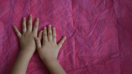 Toddler hands on crumpled tissue paper on table. Directly above view of magenta wrinkled paper and kid palms of hands touch uneven pink paper textureの写真素材