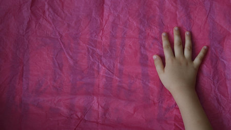 Toddlers hand on crumpled tissue paper surface. Directly above view of magenta paper and kid palm of hand touch uneven pink paper textureの写真素材