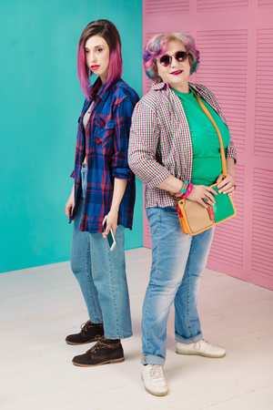 Young girl and happy senior woman in colorful casual clothes. Two generations of hipsters. Stylish senior woman on pink and blue background. Communication and common style.の写真素材