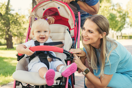 Young mother talking to smiling baby in pink stroller. Parents walking outdoors with child in summer pram.の写真素材