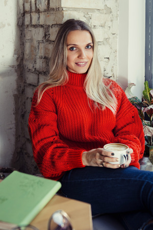 Young blonde woman sitting at a cafe window having morning latte over sunny cityの写真素材