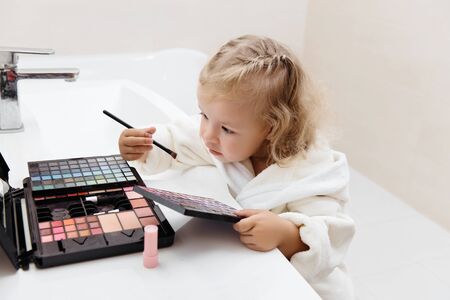 Toddler girl doing herself makeup with mamas cosmetics pretending grownup. Applying eyeshadows. Child in the bathrobe at homeの写真素材