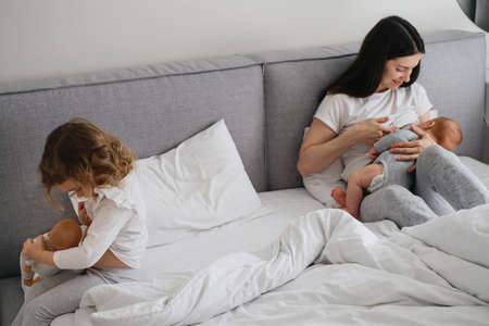 Breast feeding mama with two kids at home. Baby breastfeed on mothers hands, while older sister imitate with doll near, Cuddle a babyの写真素材