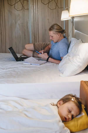 Father working while child is sleeping on the bed in hotel. Man on business trip with family.の写真素材