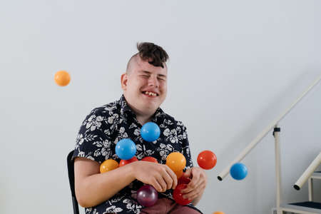 Happy teenage child over white wall in rehabilitation center. Boy with cerebral palsy smiling, playing with balls. Rehabilitation and inclusion kindergarten, day care. Disabled kid, positive emotionsの写真素材
